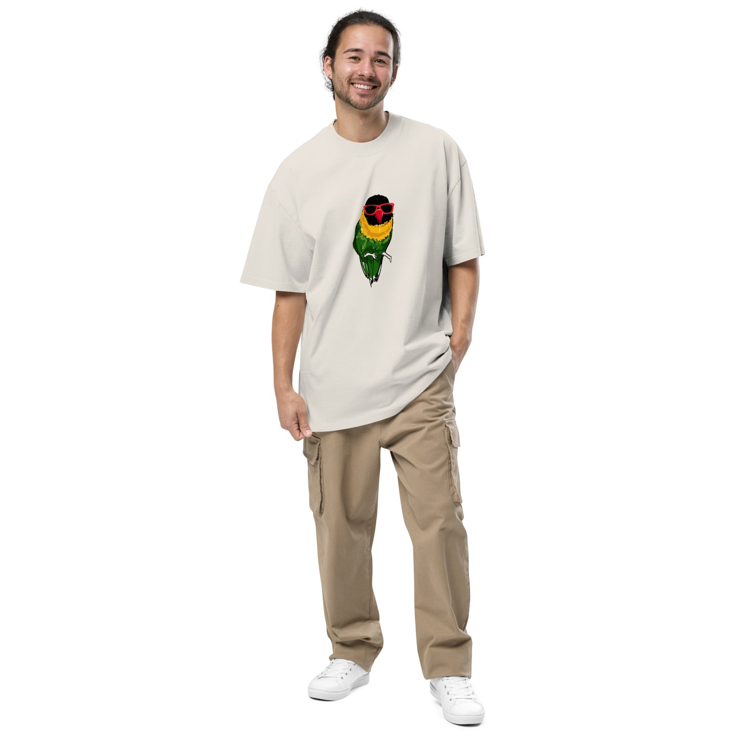 A man with a warm smile is featured wearing a stone t-shirt with a colorful graphic of a parrot wearing red sunglasses on the chest. The fun, quirky graphic adds a pop of color and humor to the outfit, which is completed with beige cargo pants and white sneakers for a comfortable, casual look.