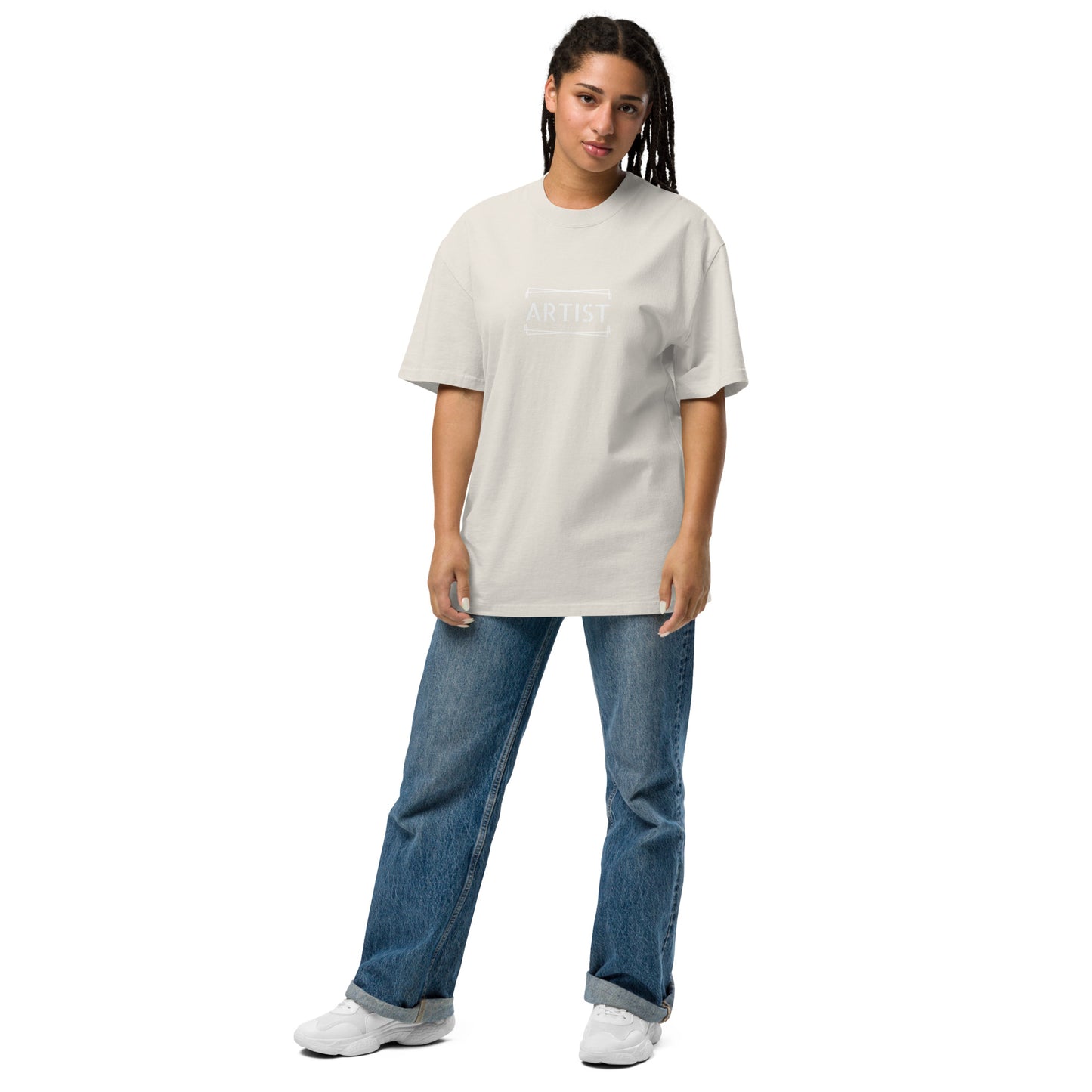 A creative individual stands confidently in a minimalist Women’s ‘ARTIST’ Statement Oversized faded bone Tee in black, paired with classic blue jeans and white sneakers. The shirt is part of a versatile graphic tee collection designed for self-expression.