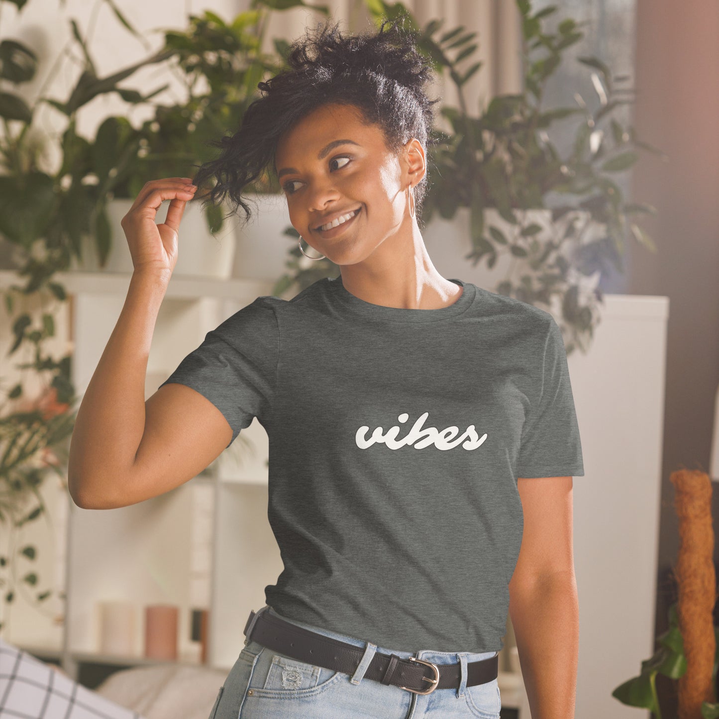 A woman with a playful touch to her hair smiles gently while wearing a heathered grey t-shirt that reads "vibes" in cursive white lettering, suggesting a laid-back, positive attitude. She's surrounded by an inviting, plant-filled indoor space.
