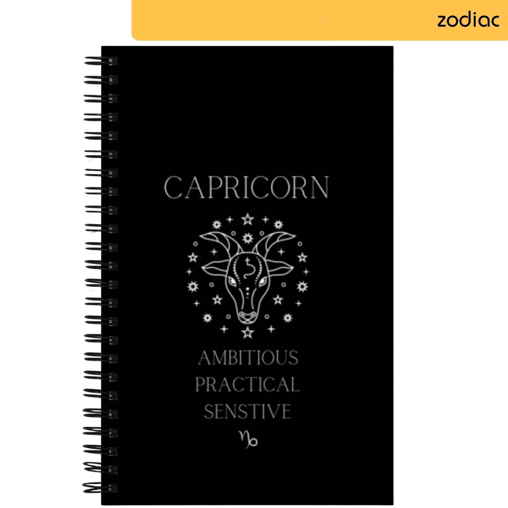 A notepad displays 'CAPRICORN' at the top in elegant white lettering. Centered below is a graphic of a goat with stars, representing the Capricorn zodiac sign. Characteristics 'AMBITIOUS PRACTICAL SENSITIVE' are listed beneath the illustration, with the Capricorn astrological symbol at the very bottom.