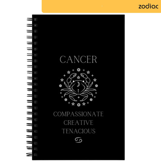 On the notepad, 'CANCER' is written in white lettering on a black background, with an artistic depiction of a crab encircled by stars above it. Listed below are traits commonly associated with Cancer: 'COMPASSIONATE CREATIVE TENACIOUS', finished with the astrological symbol for Cancer at the bottom.