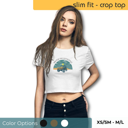 Woman in a white slim fit crop top with 'In for the Adventure' vintage truck print, paired with black high-waisted jeans, showcasing size range XS/SM to M/L and color options.