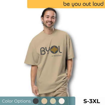 A young man wearing a tan "BYOL: a mindset" T-shirt, smiling and standing with one hand in his pocket. The T-shirt features the "BYOL" logo in dark gray and yellow on the front and is available in various colors (dark gray, beige, light blue) and sizes (S-3XL). The "be you out loud" logo is displayed at the top.