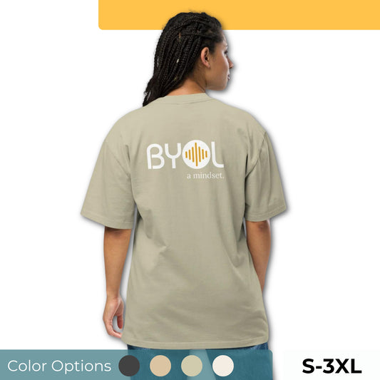 A young woman with long braids wearing a beige "BYOL: a mindset" T-shirt, viewed from the back. The T-shirt features the "BYOL" logo in white and yellow on the back and is available in multiple colors (dark gray, beige, light blue, white, plus more) and sizes (S-3XL). The "be you out loud" logo is displayed at the top.