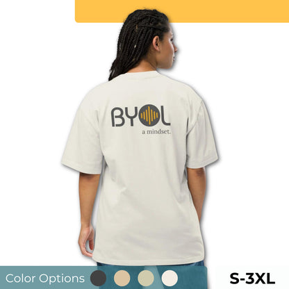 A young woman with long braids wearing a light gray "BYOL: a mindset" T-shirt, viewed from the back. The T-shirt features the "BYOL" logo in dark gray and yellow on the back and is available in multiple colors (dark gray, beige, light blue, white, plus more) and sizes (S-3XL). The "be you out loud" logo is displayed at the top.