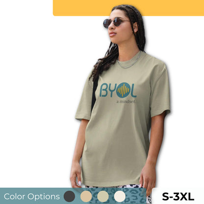 A young woman with long braids wearing sunglasses and a light eucalyptus "BYOL: a mindset" T-shirt, standing and looking to the side. The T-shirt features the "BYOL" logo in teal and yellow on the front and is available in multiple colors (dark gray, beige, light blue, white, plus more) and sizes (S-3XL). The "be you out loud" logo is displayed at the top.