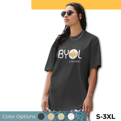 A young woman with long braids wearing sunglasses and a dark gray "BYOL: a mindset" T-shirt, standing and looking to the side. The T-shirt features the "BYOL" logo in white and yellow on the front and is available in multiple colors (dark gray, beige, light blue, white, plus more) and sizes (S-3XL). The "be you out loud" logo is displayed at the top.