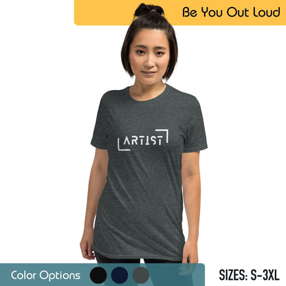 A woman with her hair in a top knot wearing a dark grey T-shirt with the word "ARTIST" printed on it, in front of a background featuring the text "Be You Out Loud" and a color options palette, with available sizes from S to 3XL noted.