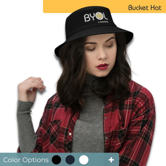 Woman wearing a black bucket hat with "BYOL a mindset" embroidered on the front in white and yellow, posing against a clean background. Available in various colors: black, dark blue, light blue, and teal.