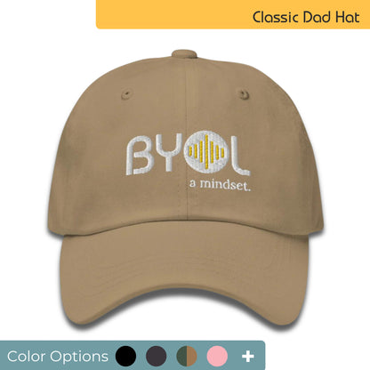 Khaki classic dad hat with "BYOL a mindset" embroidered on the front in white and yellow, displayed against a clean background. Available in various colors: black, gray, green, pink, and tan.