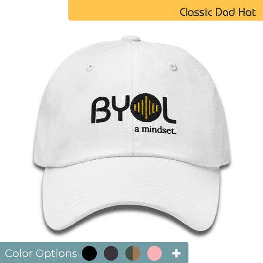 White classic dad hat with "BYOL a mindset" embroidered on the front in black and yellow, displayed against a clean background. Available in various colors: black, gray, green, pink, and white.