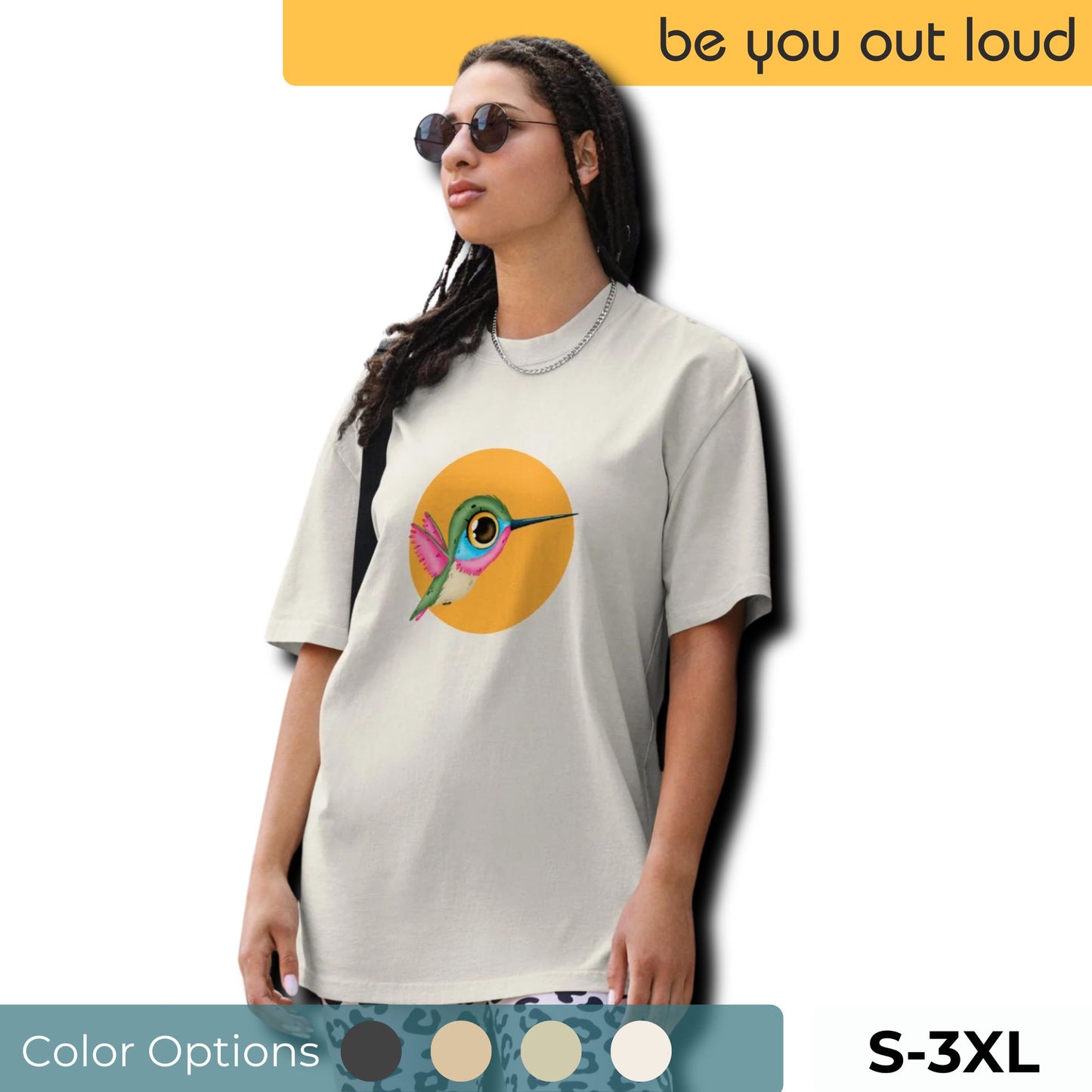 Woman models a Be You Out Loud faded bone Oversized Tee with a colorful hummingbird design on a yellow circle background, embodying a lively spirit. Shirt available with color options and sizes ranging from S to 3XL.