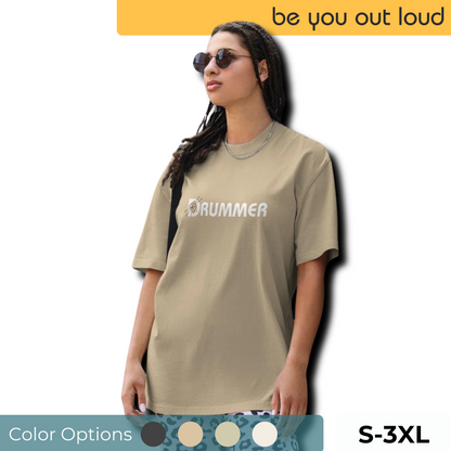 Fashion-forward woman in a minimalist faded khaki 'DRUMMER' print t-shirt, showcasing versatile color options and available from S to 3XL, exuding confidence with the 'be you out loud' mantra