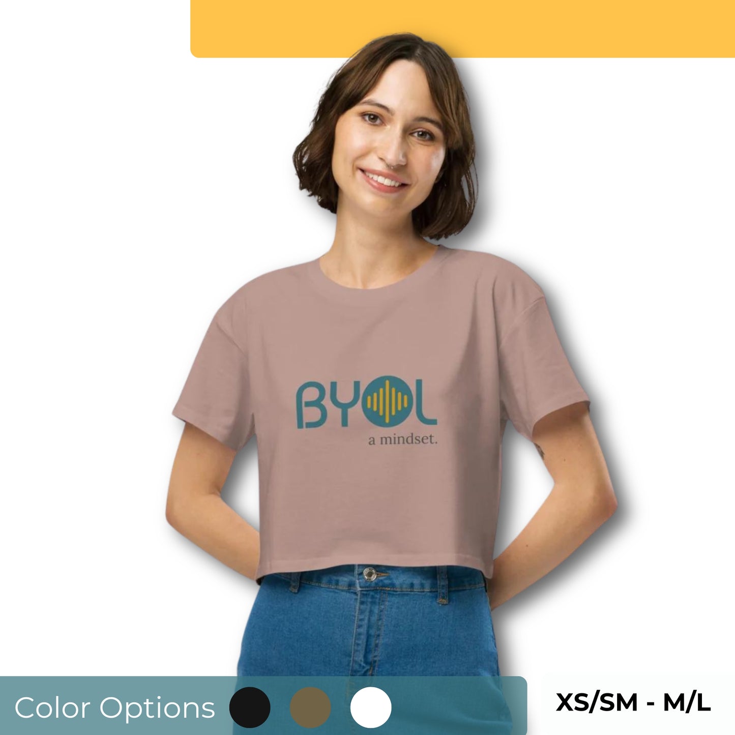 A young woman with short brown hair wearing a hazy pink "BYOL: a mindset" crop top, smiling and standing with her hands on her hips. The crop top features the "BYOL" logo in teal and yellow on the front and is available in multiple colors (black, brown, white, light blue, plus more) and sizes (XS/SM - M/L). The "be you out loud" logo is displayed at the top.