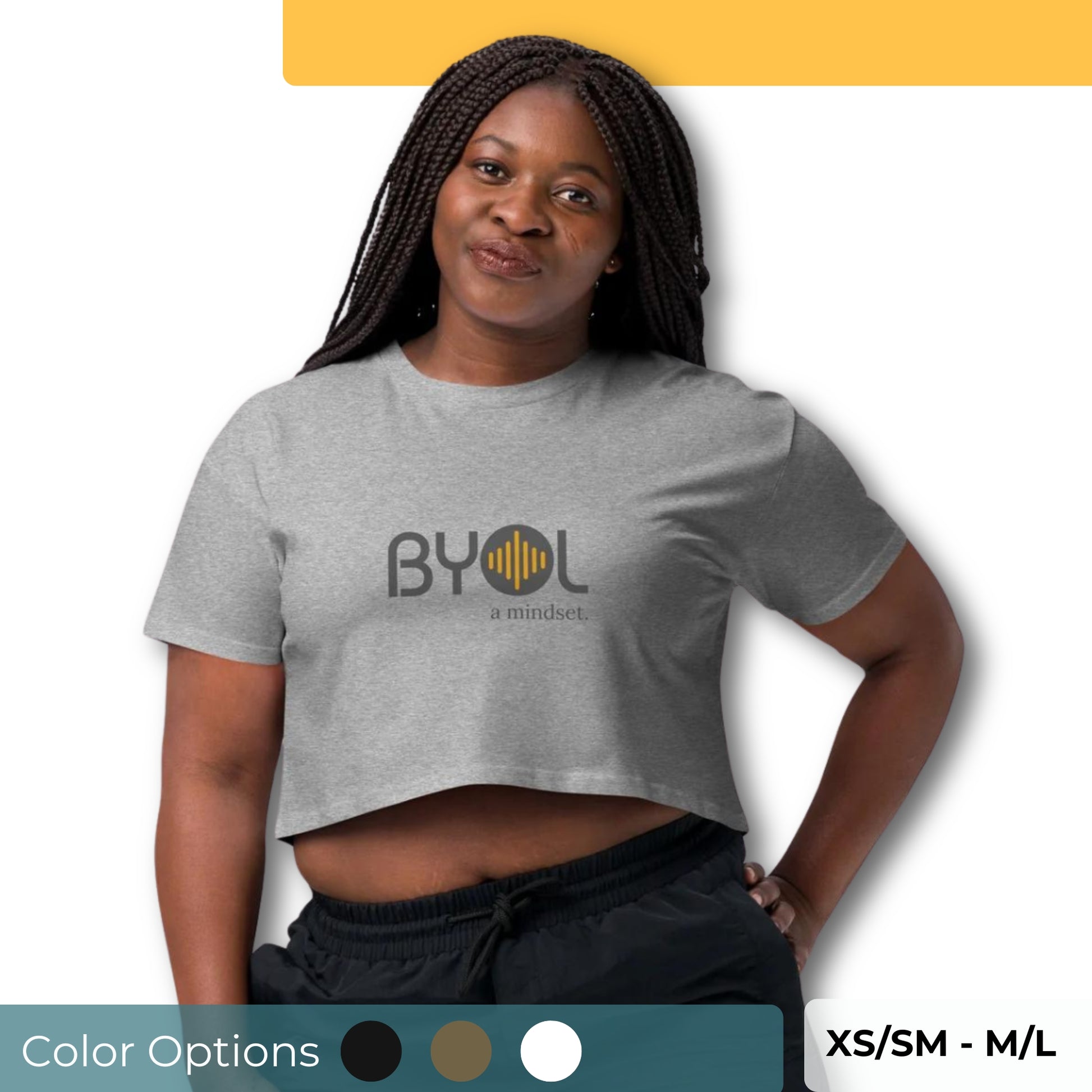 A young woman with long braided hair wearing a gray "BYOL: a mindset" crop top, standing with a hand on her hip and slightly pouting. The crop top features the "BYOL" logo in black and yellow on the front and is available in multiple colors (black, brown, white, light blue, plus more) and sizes (XS/SM - M/L). The "be you out loud" logo is displayed at the top.