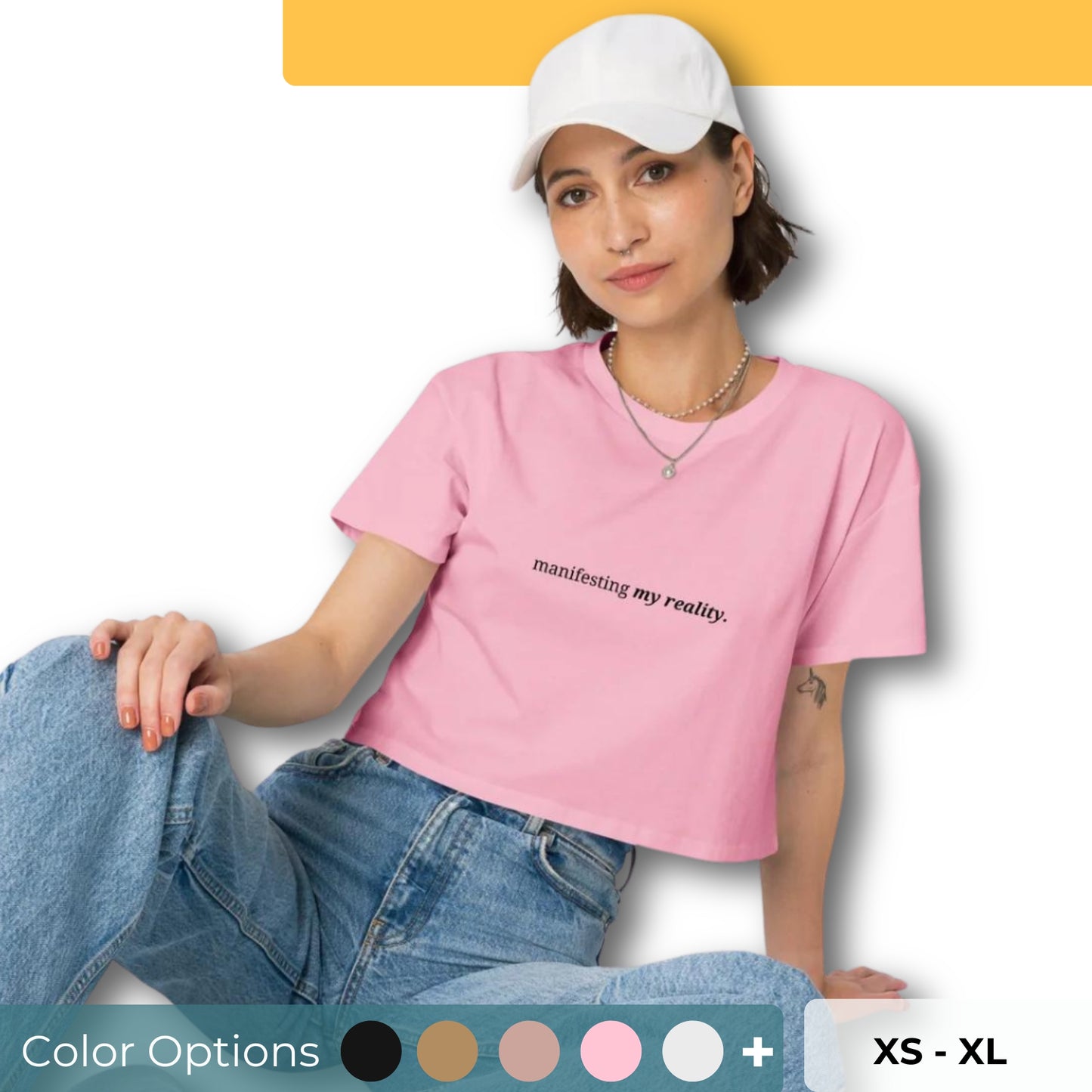 Woman wearing a bubblegum pink cropped t-shirt with the phrase "manifesting my reality" printed in black, sitting and posing against a clean background. Available in various colors: black, brown, pink, green, and white. Sizes range from XS to XL.