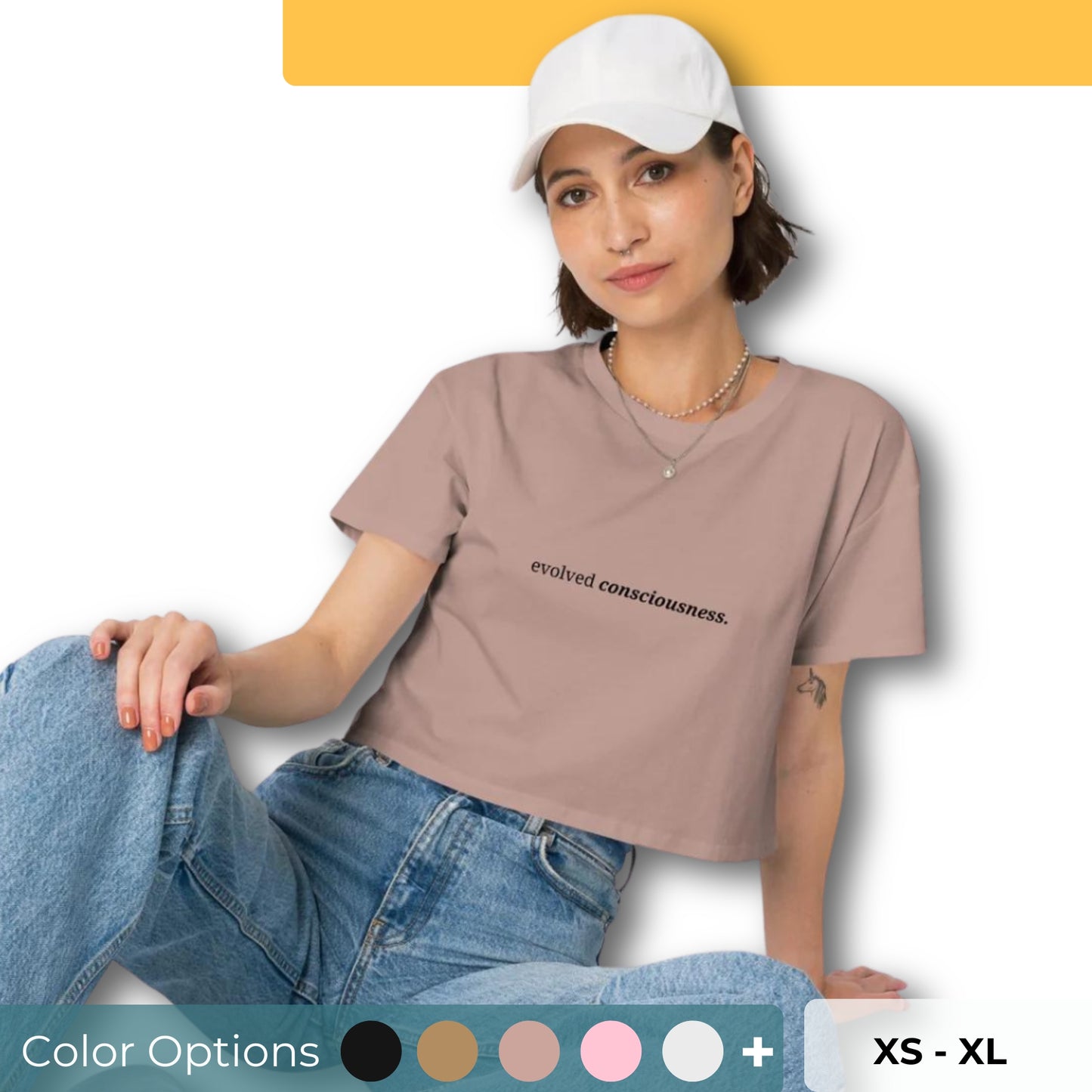 Woman wearing a hazy pink cropped t-shirt with the phrase "evolved consciousness" printed in black, sitting and posing against a clean background. Available in various colors: black, brown, pink, green, and white. Sizes range from XS to XL.