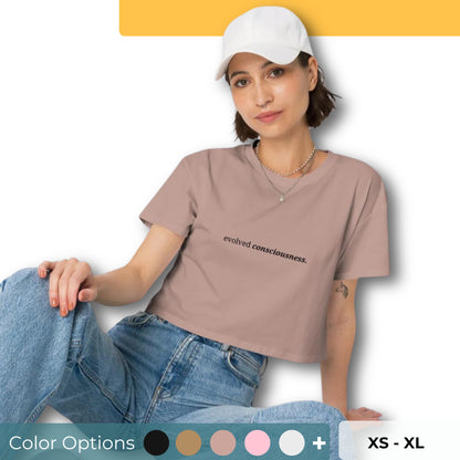 Woman wearing a hazy pink cropped t-shirt with the phrase "evolved consciousness" printed in black, sitting and posing against a clean background. Available in various colors: black, brown, pink, green, and white. Sizes range from XS to XL.