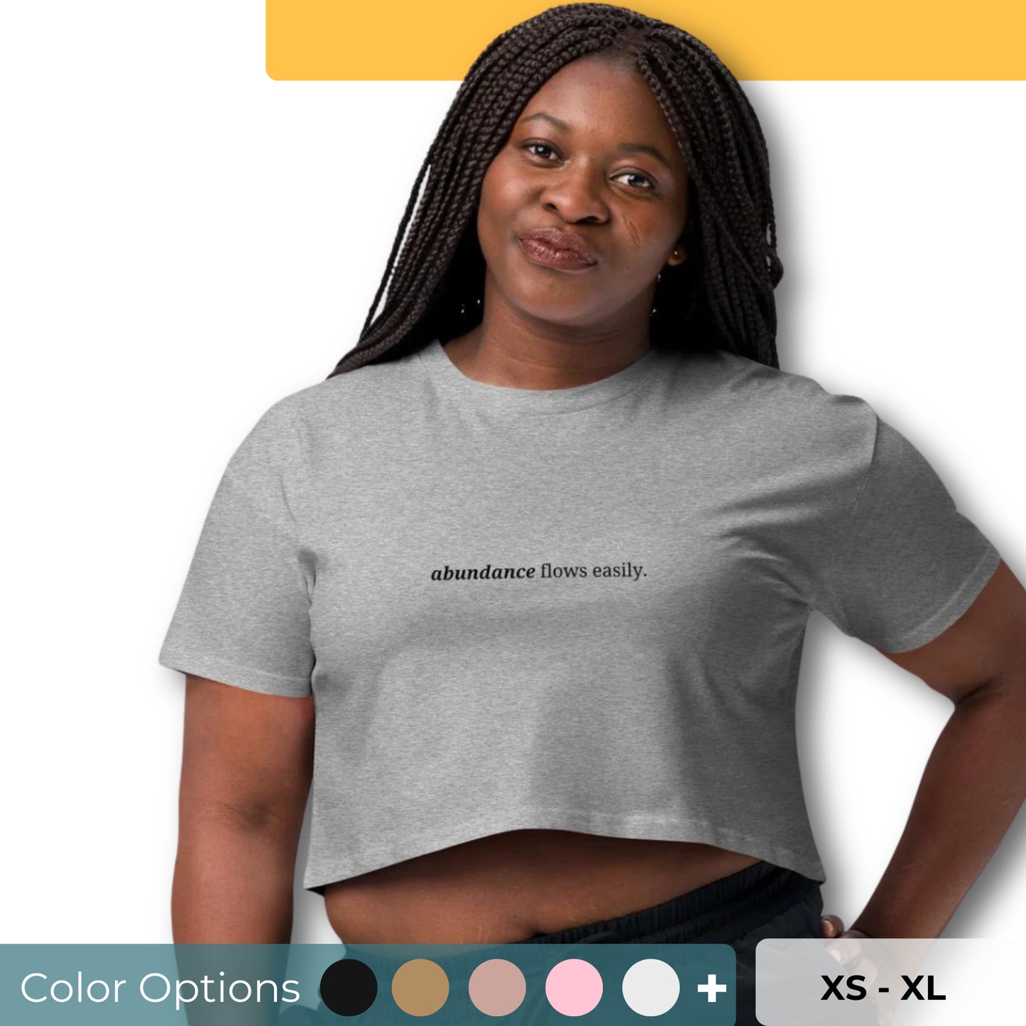 Woman wearing a heather gray cropped t-shirt with the phrase "abundance flows easily" printed in black, smiling and posing against a clean background. Available in various colors: black, brown, pink, green, and white. Sizes range from XS to XL.