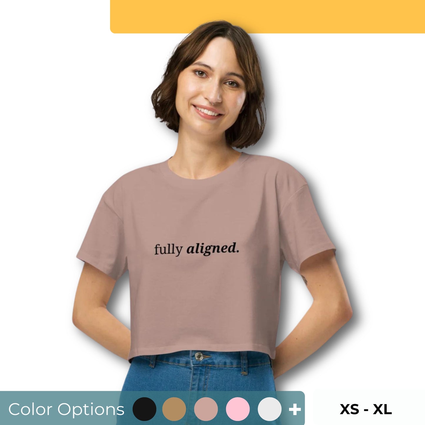 Woman wearing a hazy pink cropped t-shirt with the phrase "fully aligned" printed in black, smiling and posing against a clean background. Available in various colors: black, brown, pink, green, and white. Sizes range from XS to XL.