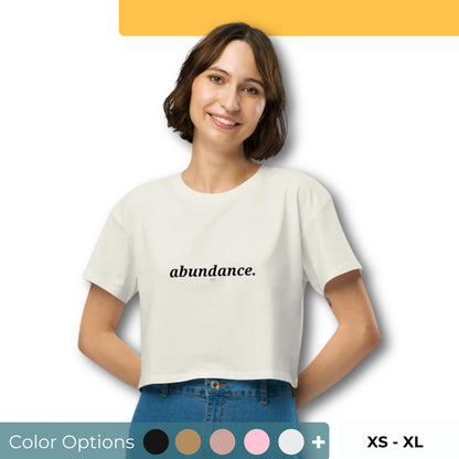 Woman wearing a ecru cropped t-shirt with the word "abundance" printed in black, smiling, and posing against a clean background. Available in various colors: black, brown, pink, green, and white. Sizes range from XS to XL.