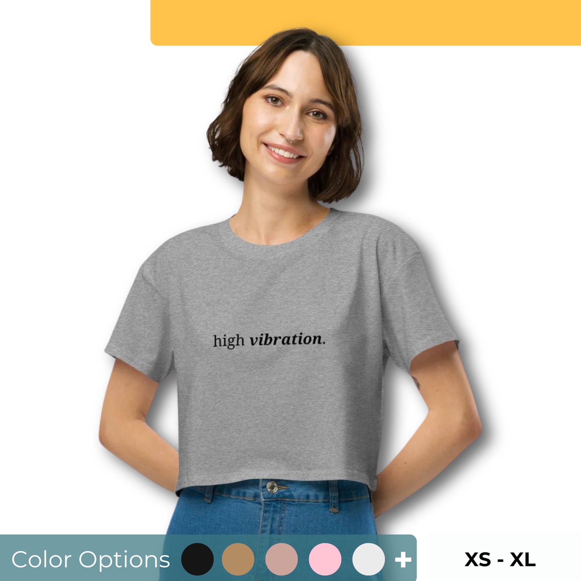 Woman wearing a heather gray cropped t-shirt with the phrase "high vibration" printed in black, smiling and posing against a clean background. Available in various colors: black, brown, pink, green, and white. Sizes range from XS to XL.