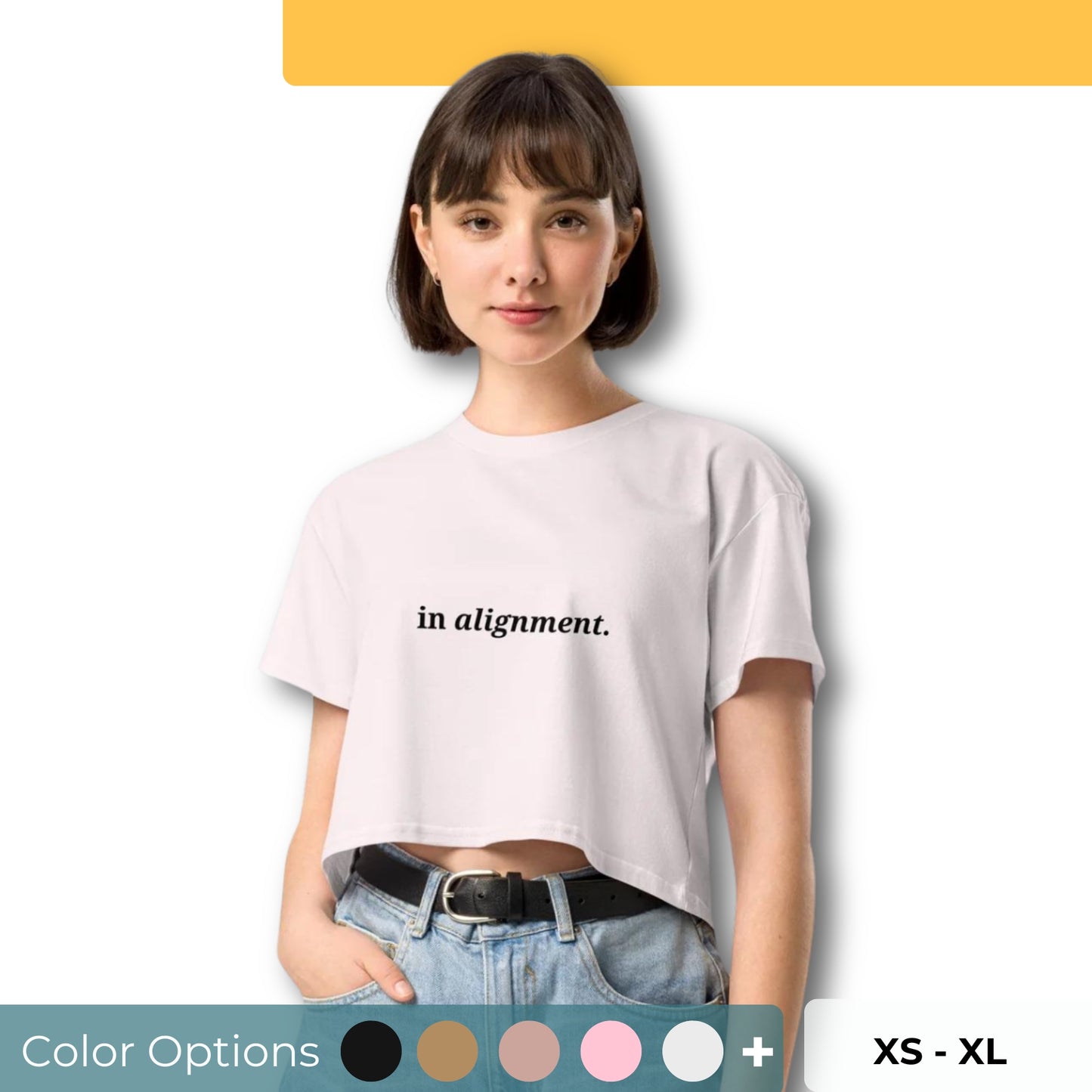 Woman wearing a orchid cropped t-shirt with the phrase "in alignment" printed in black, posing against a clean background. Available in various colors: black, brown, pink, green, and white. Sizes range from XS to XL.