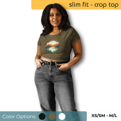 Confident woman wearing a slim fit olive green crop top with a colorful mountain sunset design, paired with high-waisted grey jeans, available in sizes XS/SM to M/L.