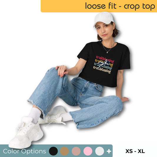 Casual woman sitting with one knee up, wearing a black loose-fit crop top with 'trailblazing' text, light-wash denim jeans, white sneakers, and a white cap, with color options from XS to XL visible.