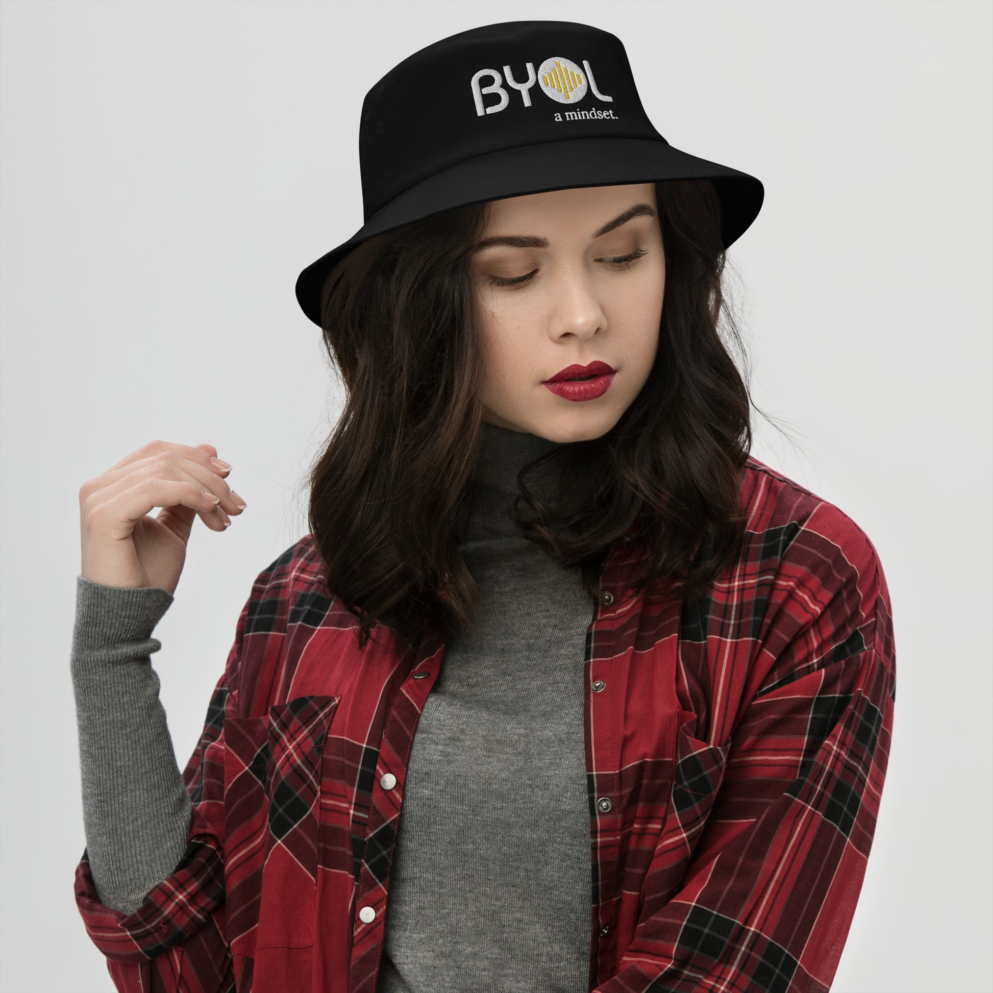 Woman wearing a black bucket hat with "BYOL a mindset" embroidered on the front in white and yellow, posing against a clean background. Available in various colors: black, dark blue, light blue, and teal.