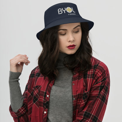 Woman wearing a navy bucket hat with "BYOL a mindset" embroidered on the front in white and yellow, posing against a clean background. Available in various colors: black, dark blue, light blue, and teal.