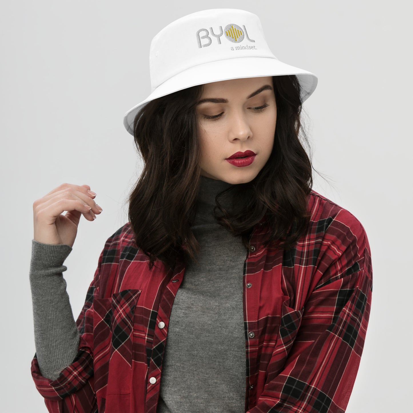 Woman wearing a white bucket hat with "BYOL a mindset" embroidered on the front in white and yellow, posing against a clean background. Available in various colors: black, dark blue, light blue, and teal.