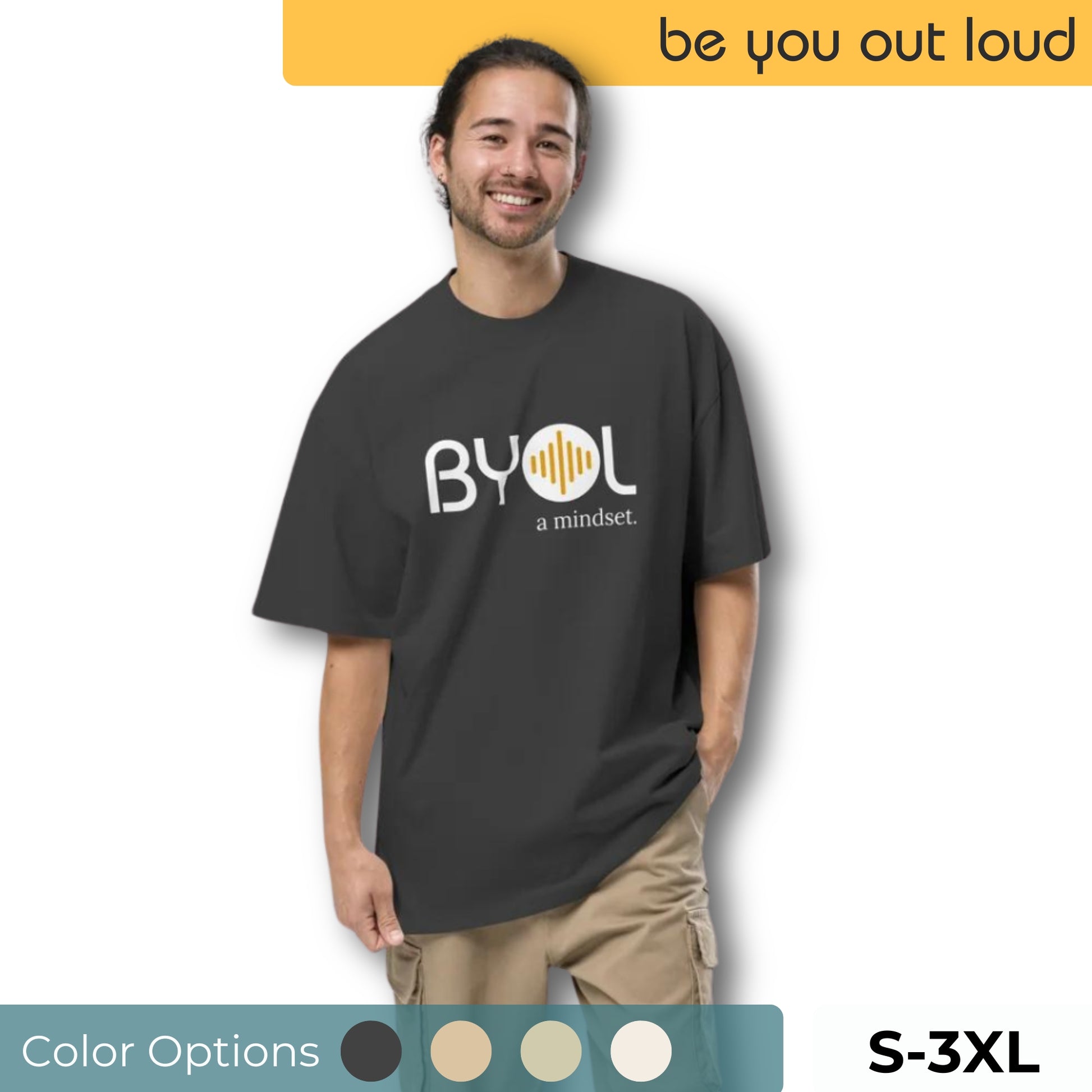 A young man wearing a dark gray "BYOL: a mindset" T-shirt, smiling and standing with one hand in his pocket. The T-shirt features the "BYOL" logo in white and yellow on the front and is available in various colors (dark gray, beige, light blue) and sizes (S-3XL). The "be you out loud" logo is displayed at the top.