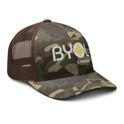 Side of camo/brown retro trucker hat with "BYOL a mindset" embroidered on the front in white and yellow, featuring a black mesh back, displayed against a clean background. Available in various colors: black, brown, green, and beige.