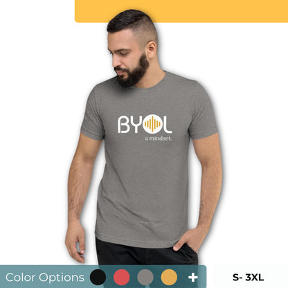 A young man with short dark hair and a beard wearing a gray "BYOL: a mindset" T-shirt, standing with one hand in his pocket and looking down. The T-shirt features the "BYOL" logo in white and yellow on the front and is available in multiple colors (black, red, gray, pink, gold, plus more) and sizes (S-3XL). The "be you out loud" logo is displayed at the top.