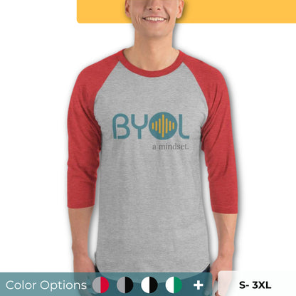 A young man wearing a gray and red "BYOL: a mindset" raglan T-shirt with three-quarter sleeves, smiling and standing with his arms at his sides. The T-shirt features the "BYOL" logo in teal and yellow on the front and is available in multiple colors (red, black, white, green, plus more) and sizes (S-3XL). The "be you out loud" logo is displayed at the top.