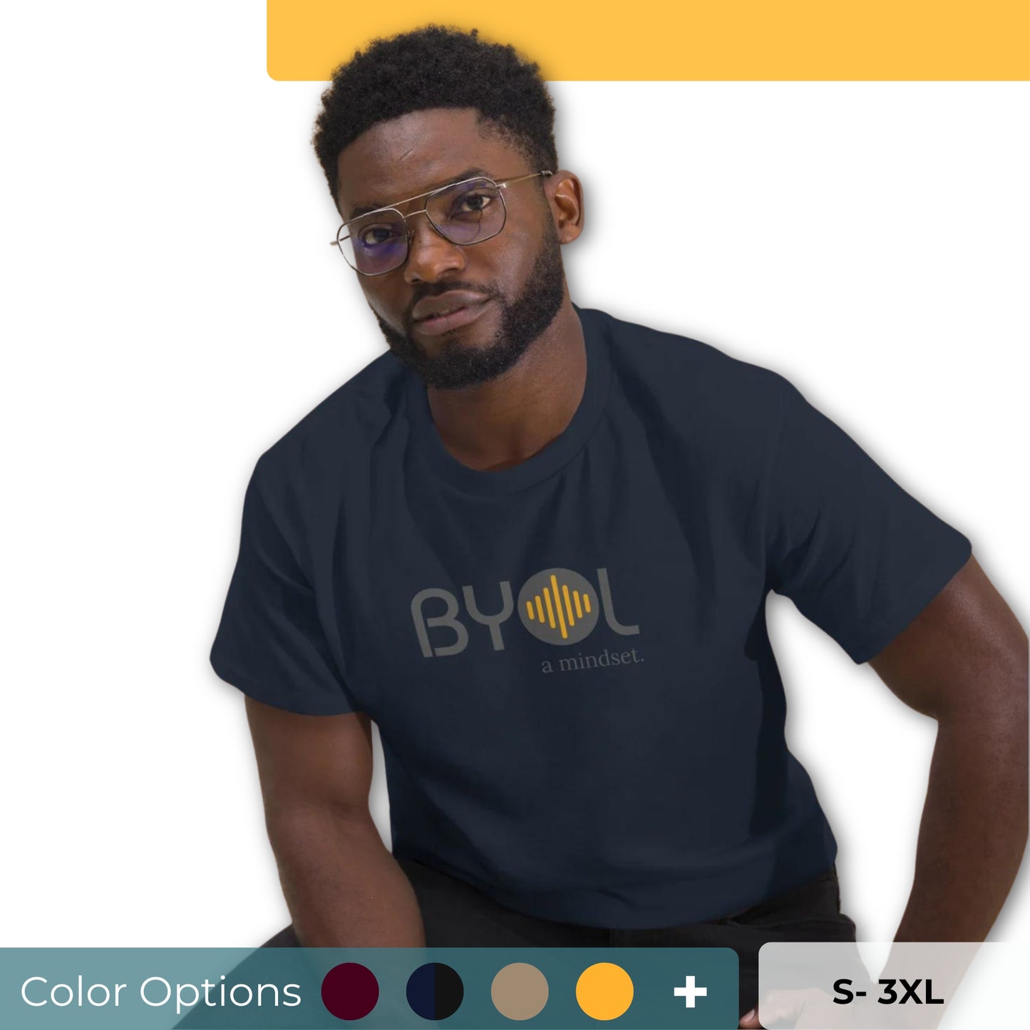 A young man with short curly hair and glasses wearing a dark blue "BYOL: a mindset" T-shirt, sitting down. The T-shirt features the "BYOL" logo in gray and yellow on the front and is available in multiple colors (maroon, black, brown, gold, plus more) and sizes (S-3XL). The "be you out loud" logo is displayed at the top.