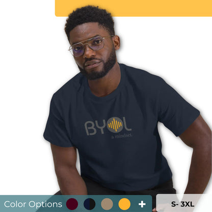 A young man with short curly hair and glasses wearing a dark blue "BYOL: a mindset" T-shirt, sitting down. The T-shirt features the "BYOL" logo in gray and yellow on the front and is available in multiple colors (maroon, black, brown, gold, plus more) and sizes (S-3XL). The "be you out loud" logo is displayed at the top.