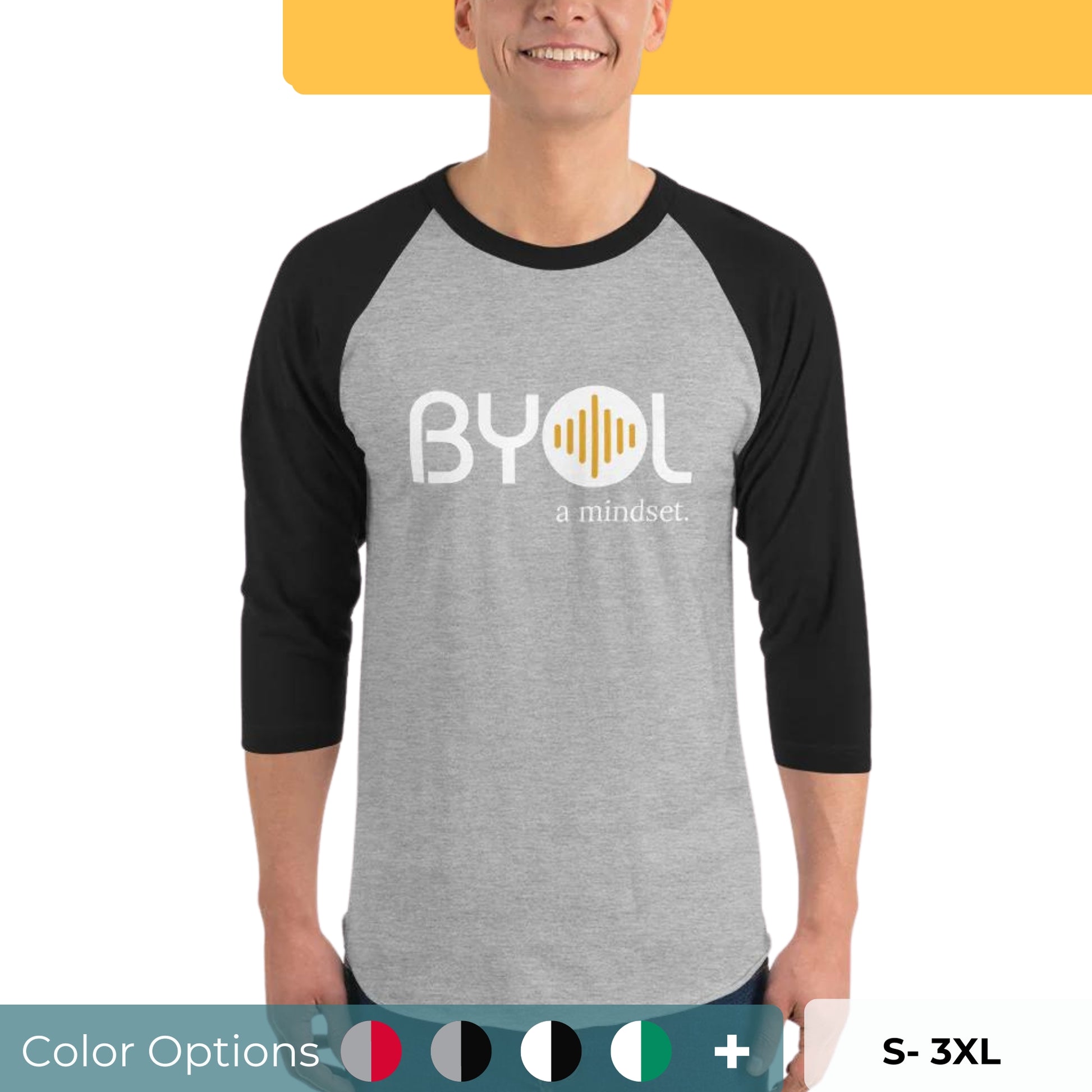 A young man wearing a gray and black "BYOL: a mindset" raglan T-shirt with three-quarter sleeves, smiling and standing with his arms at his sides. The T-shirt features the "BYOL" logo in white and yellow on the front and is available in multiple colors (red, black, white, green, plus more) and sizes (S-3XL). The "be you out loud" logo is displayed at the top.