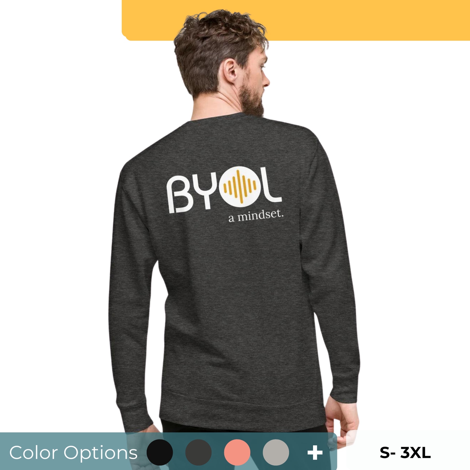 A young man with short hair and a beard wearing a dark gray "BYOL: a mindset" sweatshirt, viewed from the back. The sweatshirt features the "BYOL" logo in white and yellow on the back and is available in multiple colors (black, dark gray, light gray, pink, plus more) and sizes (S-3XL). The "be you out loud" logo is displayed at the top.