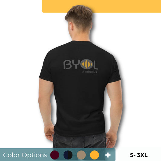 A young man with short blonde hair wearing a black "BYOL: a mindset" T-shirt, viewed from the back. The T-shirt features the "BYOL" logo in gray and yellow on the back and is available in multiple colors (maroon, black, brown, gold, plus more) and sizes (S-3XL). The "be you out loud" logo is displayed at the top.