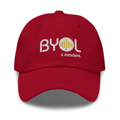 Red classic dad hat with "BYOL a mindset" embroidered on the front in white and yellow, displayed against a clean background. Available in various colors: black, gray, green, pink, and tan.