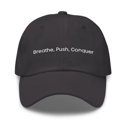 A dark grey dad hat with the phrase "Breath,  Push, Conquer" embroidered in white stitching across the front