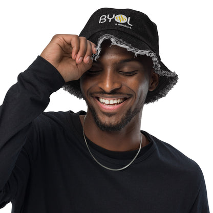 Man wearing a black bucket hat with frayed edges and "BYOL a mindset" embroidered on the front in white and yellow, smiling and posing against a clean background. Available in various colors: black, dark blue, light blue, and teal.