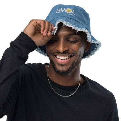 Man wearing a light denim bucket hat with frayed edges and "BYOL a mindset" embroidered on the front in white and yellow, smiling and posing against a clean background. Available in various colors: black, dark blue, light blue, and teal.