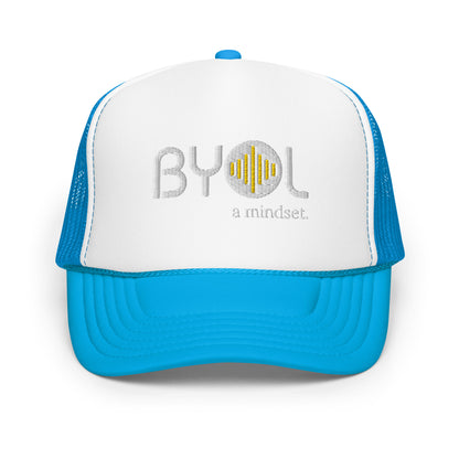 White and blue foam trucker hat with "BYOL a mindset" embroidered on the front in gray and yellow, displayed against a clean background. Available in various colors: black, white and blue, blue, pink, and white.