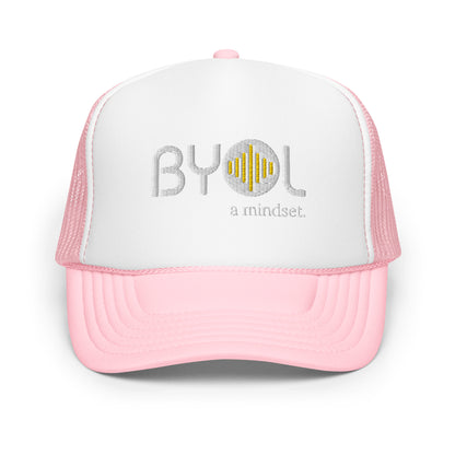 White and pink foam trucker hat with "BYOL a mindset" embroidered on the front in gray and yellow, displayed against a clean background. Available in various colors: black, white and blue, blue, pink, and white.