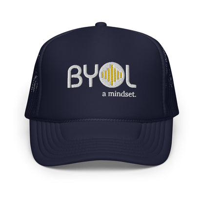 Blue on blue foam trucker hat with "BYOL a mindset" embroidered on the front in gray and yellow, displayed against a clean background. Available in various colors: black, white and blue, blue, pink, and white.