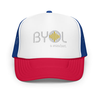 White, red, and blue foam trucker hat with "BYOL a mindset" embroidered on the front in gray and yellow, displayed against a clean background. Available in various colors: black, white and blue, blue, pink, and white.