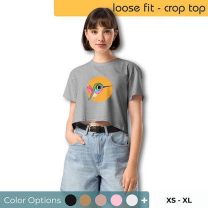 Woman in a grey loose-fit crop top with a vibrant eye and hummingbird graphic, available in multiple colors and sizes XS through XL.